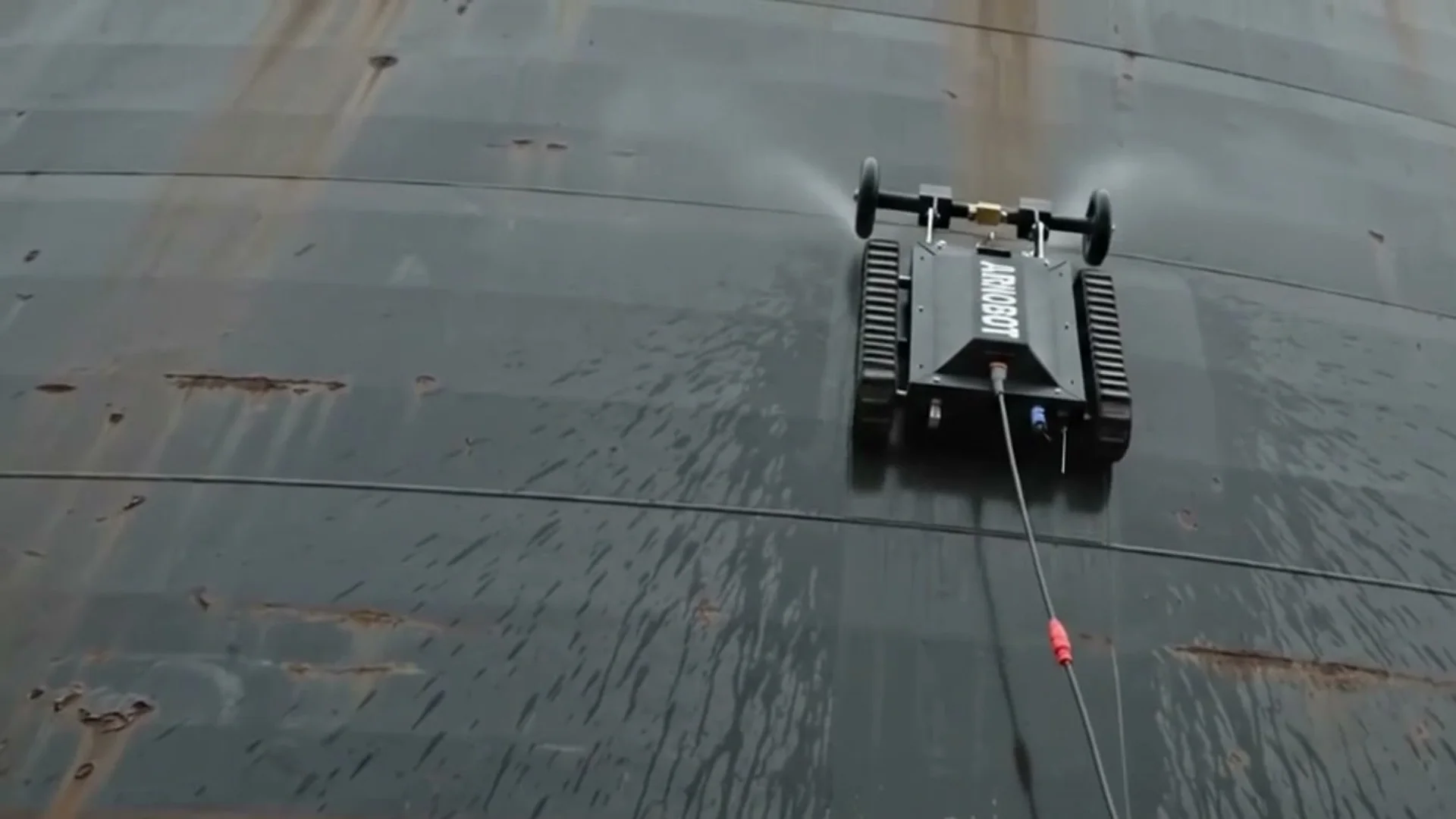 ALTIUS climbing a wall of dark ship-hull plating, its spray bar running and its tether trailing down the steel