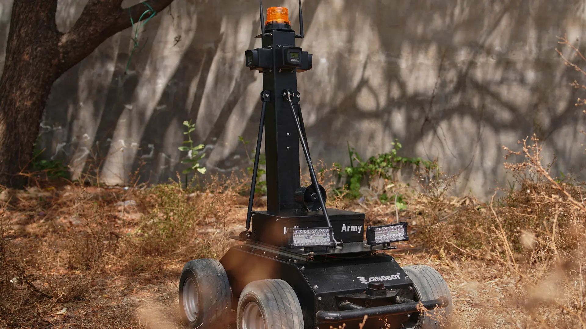 Saibya standing in dry scrub against a shaded wall on a field trial, sensor mast and beacon up, light bars across its deck